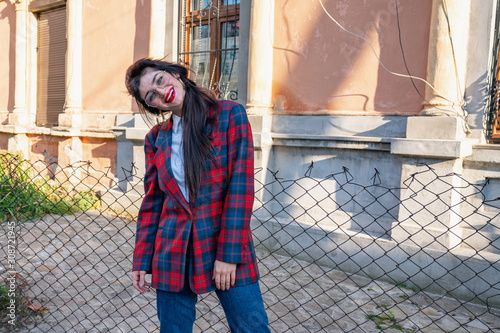 Attaractive brunette girl in glasses dressed in plaid jacket and white shirt bowed her head on left shoulder and looking at the camera stands near the old house photo