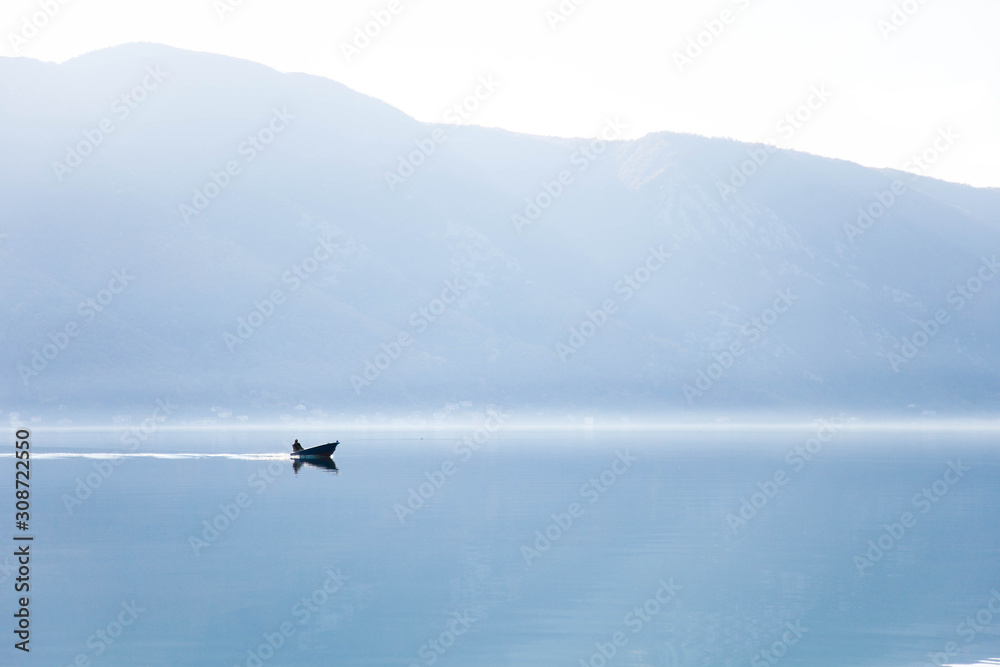 Fototapeta premium Blue background. Boat with fisherman on sea. Fishing in foggy morning lake. Amazing nature landscape with mountains, silence, calmness. Reflection in still water of Kotor Bay, Montenegro. Copy space