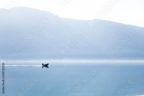 Blue background. Boat with fisherman on sea. Fishing in foggy morning lake. Amazing nature landscape with mountains, silence, calmness. Reflection in still water of Kotor Bay, Montenegro. Copy space