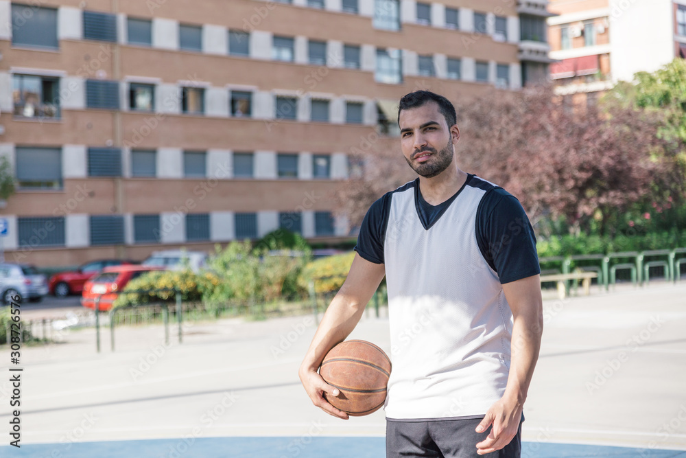 Strong and attractive basketball player looks at camera smiling while posing with his ball on an urban court Strong and attractive basketball player looks at camera smiling while posing with his ball on an urban court