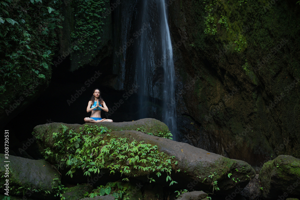 Young Caucasian woman meditating, practicing yoga at waterfall. Hands in namaste mudra. Leke Leke waterfall, Bali, Indonesia.