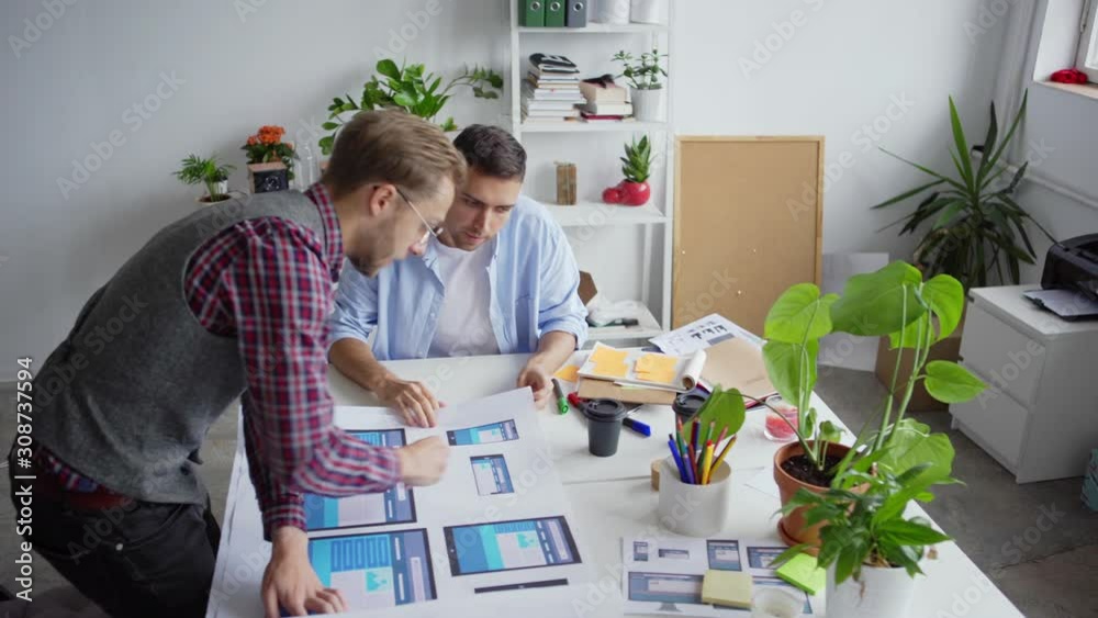 High angle view of two designers examining and discussing website interface samples for different devices printed on paper sheet at desk in office High angle view of two designers examining and discussing website interface samples for different devices printed on paper sheet at desk in office
