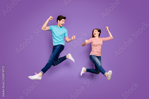 Full body photo of funny two people guy lady jumping high rejoicing ecstatic achievement raise fists wear casual blue striped t-shirts jeans footwear isolated purple color background