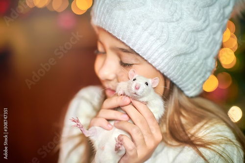 Cute little girl in a hat holds in her arms and hugs a funny white rat on a background of bright holiday lights. Symbol of 2020. Pets