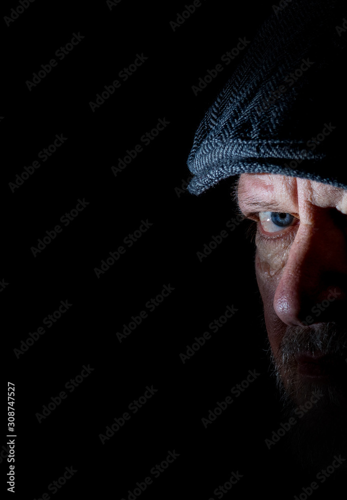 Portrait of a senior man with serious face expression. His face is wrinkled and only partially illuminated. He is wearing a flat cap. The background is black. Portrait of a senior man with serious face expression. His face is wrinkled and only partially illuminated. He is wearing a flat cap. The background is black.