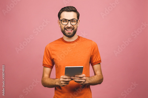 Happy young man in casual standing and using tablet isolated over pink background.