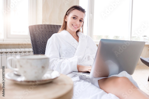 Smiling positive young girl in a white coat checks mail with laptop and wireless internet in morning after shower with a cup of coffee. Concept of the beginning of working day