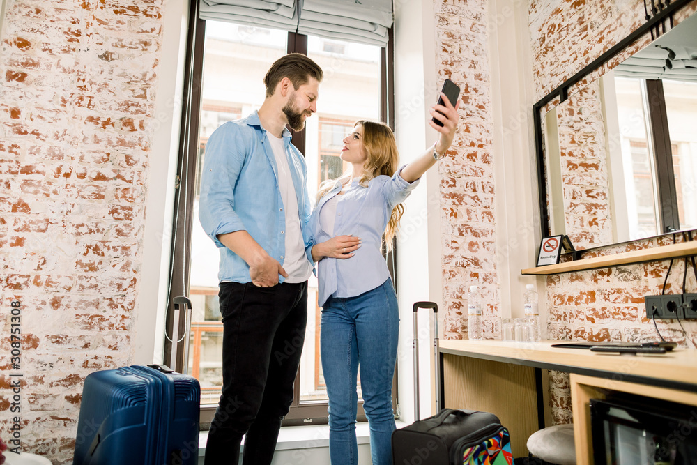 Happy young couple taking selfie with mobile phone while standing together over the window background at a hotel room with a suitcases