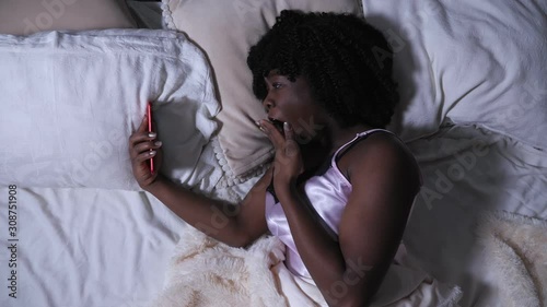 beautiful black lady looks at modern smartphone display and yawns lying on queen size bed at dark night slow motion photo