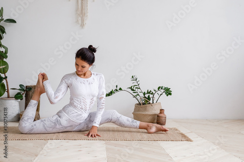 Slim pretty young brunette woman in a tight light suit makes hanumanasana sitting on a rug on the floor surrounded by houseplants on a white background. Advanced amateur concept. Advertising space photo