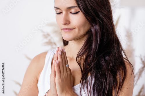 Portrait of a beautiful peaceful meditating young brunette woman with closed eyes put her hands on her chest in a cozy home atmosphere. ?oncept of relaxation and meditation. Advertising space