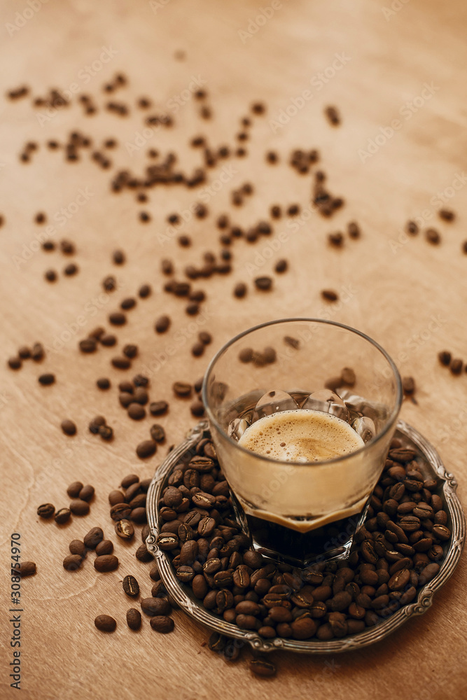 Hot espresso with foam in glass cup on vintage plate with aromatic roasted coffee beans on wooden table. Copy space. Alternative Coffee brewing concept. Hot espresso with foam in glass cup on vintage plate with aromatic roasted coffee beans on wooden table. Copy space. Alternative Coffee brewing concept.