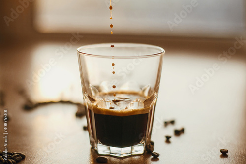 Obraz na plátně Coffee drops close up in warm light while pouring hot espresso in glass cup on background of roasted coffee beans on wooden table