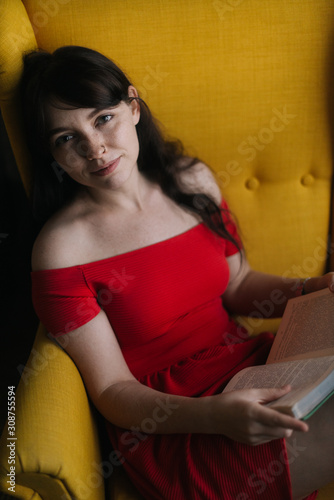 Happy young woman is reading a book in a yellow soft chair. Girl looking at the camera. Pretty girl in provocative bright clothes.