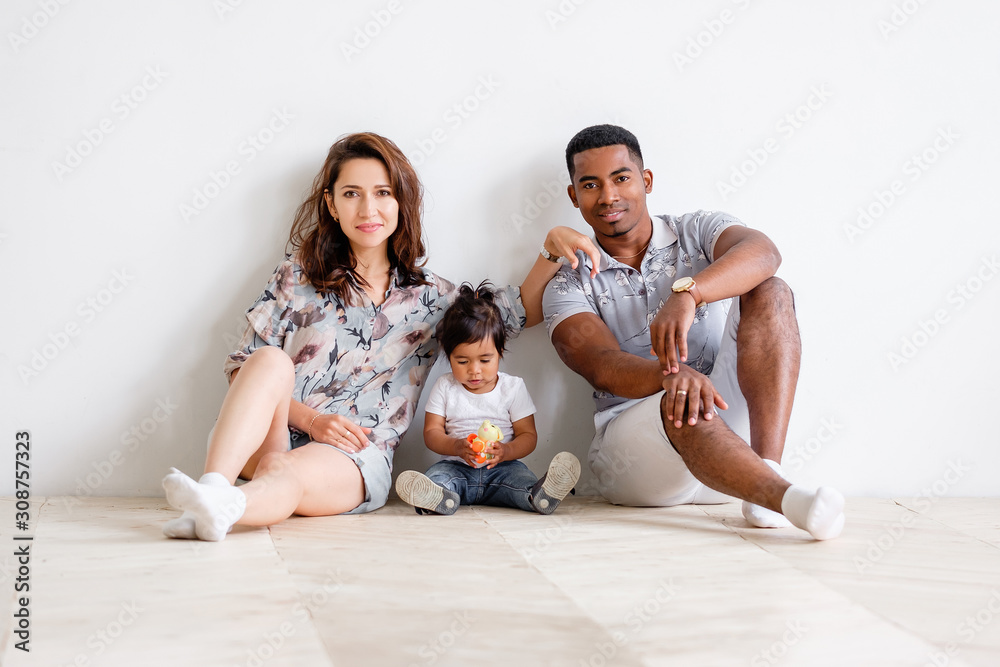 Loving young woman is looking at her young african-american husband while sitting on floor with her little charming mixed-race daughter. Concept of interracial american happy family. Advertising space