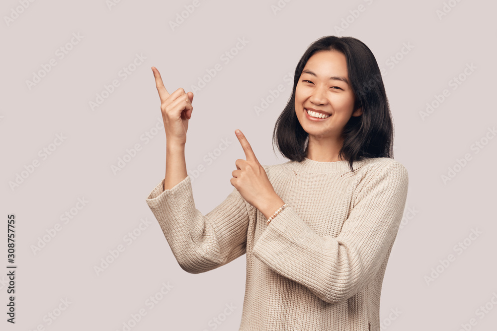 Smiling girl shows finger to the side and up. Indicate free space. Businesslike young woman Asian appearance with black hair and brown eyes dressed in knitted warm sweater stands isolated background Smiling girl shows finger to the side and up. Indicate free space. Businesslike young woman Asian appearance with black hair and brown eyes dressed in knitted warm sweater stands isolated background