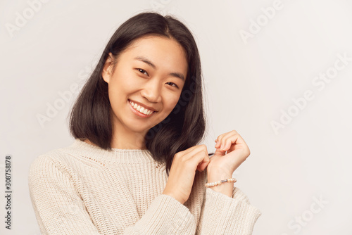 Girl nice smiles, holds hands have faces, laughs. Businesslike young woman Asian appearance with black hair and brown eyes dressed in knitted warm sweater stands isolated white background in Studio.