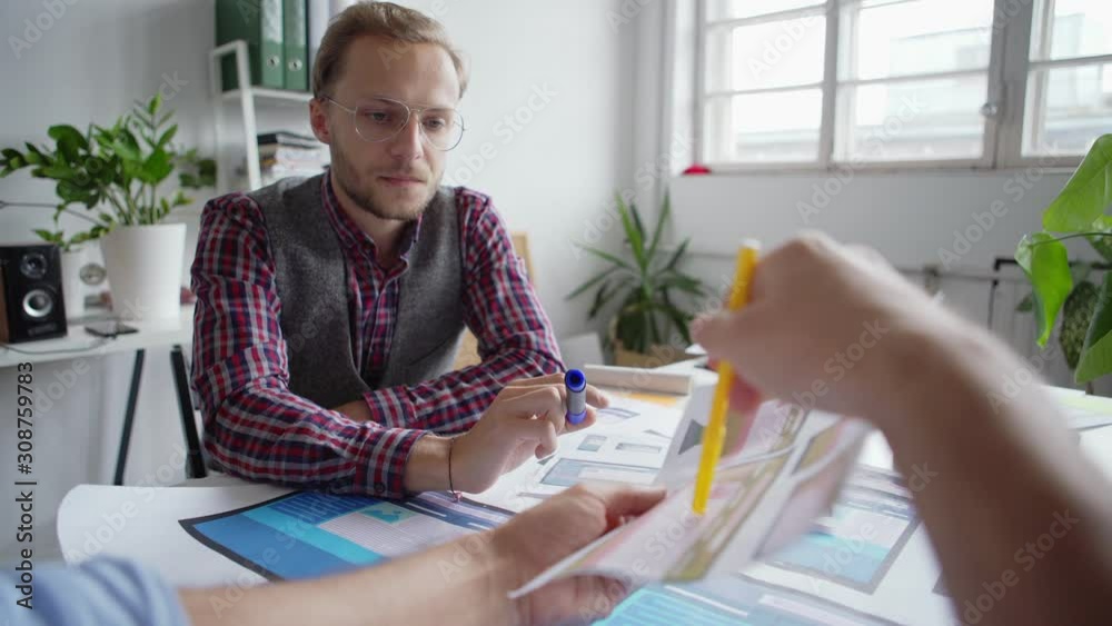 POV of young designer in eyeglasses listening to colleague showing him website interface samples for different devices printed on paper sitting at desk in office