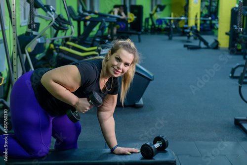 Fototapeta Naklejka Na Ścianę i Meble -  An obese woman is training with dumbbells on a bench in the gym. The fat blonde is losing weight with the help of fitness.