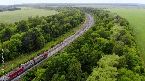 Aerial shot of freight train that travels by rail. Train carries goods and raw materials for heavy industry. Railways are surrounded by trees and endless green fields. Taken by drone at sunset