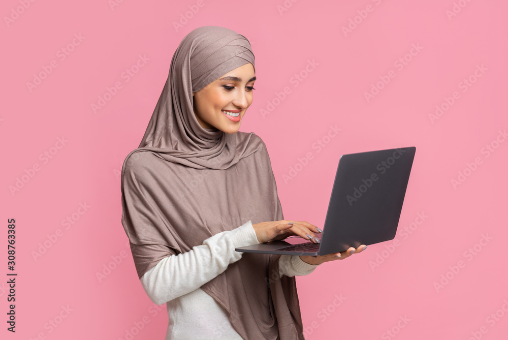 Smiling muslim girl in hijab holding and typing on laptop Smiling muslim girl in hijab holding and typing on laptop