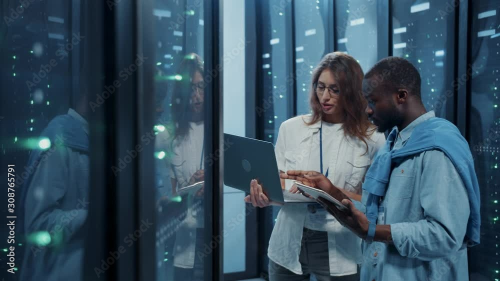 Female IT professional instructing young server technician inside working server cabinet. Male and female server engineers coworking with laptop inspecting data center cyber security.