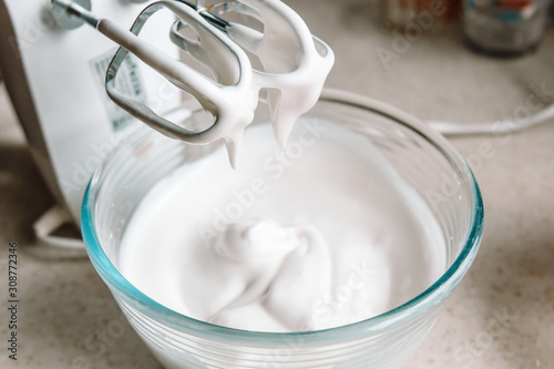 Baking concept. Whipped cream of egg whites for perfect peaks in a glass bowl, with a mixer on a marble white table. Meringue cookie recipe. photo