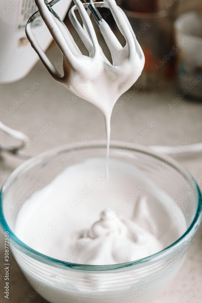 Baking concept. Whipped cream of egg whites for perfect peaks in a glass bowl, with a mixer on a marble white table. Meringue cookie recipe. Baking concept. Whipped cream of egg whites for perfect peaks in a glass bowl, with a mixer on a marble white table. Meringue cookie recipe.