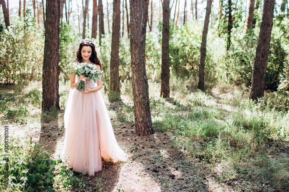 Beautiful bride in a tender pink dress outdoors. Porter in full growth of a beautiful brunette woman with long dark hair. Girl holds a bouquet. A girl in a long dress walks in a pine forest.