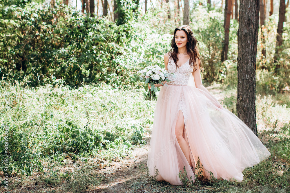 Beautiful bride in a tender pink dress outdoors. Porter in full growth of a beautiful brunette woman with long dark hair. Girl holds a bouquet. A girl in a long dress walks in a pine forest.
