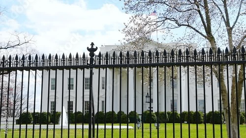 Loopable cinemagraph slow motion view through fence on White House in Washington DC, USA with fountain splashing water in spring by green grass lawn and American flag
