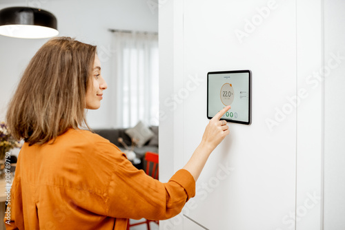 Young woman controlling temperature in the living room with a digital touch screen panel installed on the wall. Concept of heating control in a smart home photo
