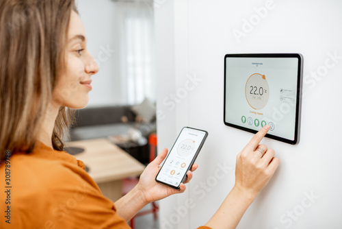 Young woman controlling temperature in the living room with smart phone and digital touch screen panel. Concept of heating control in a smart home photo