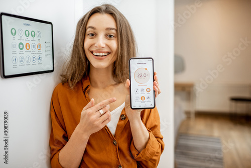 Portrait of a young and happy woman near a touch screen panel for managing smart home. Holding smart phone with a simlpe and convinient mobile application for smart home photo