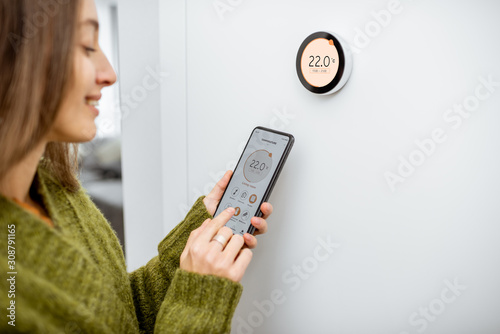 Woman dressed in green sweater regulating heating temperature with a modern wireless thermostat and smart phone at home. Synchronization of thermostat with mobile devices concept