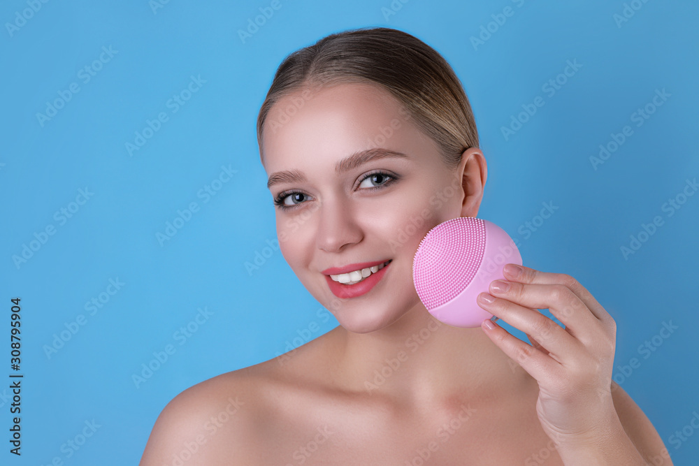 Young woman washing face with cleansing brush on light blue background. Cosmetic product Young woman washing face with cleansing brush on light blue background. Cosmetic product