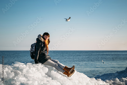 Young smiling woman sitting on a top of frozen sea ice blocks on a coast of the sea with a blue sky at the background, at frosty sunny day.