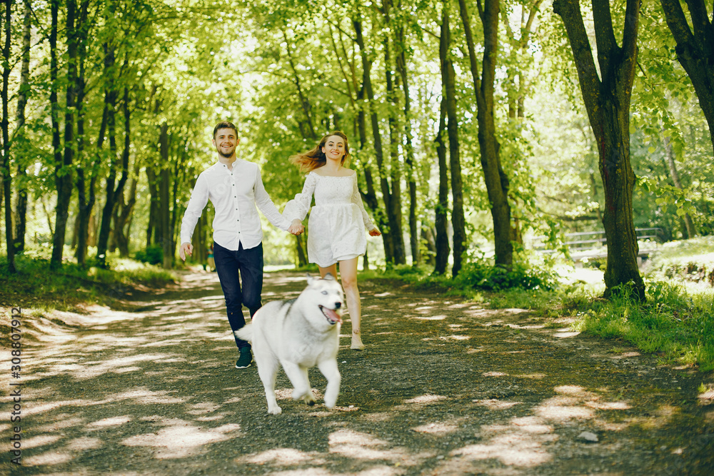 A beautiful and gentle girl with light hair and a white dress is walking in a sunny summer forest with her handsome guy in a white shirt and dark pants and they playing with cute gray dog A beautiful and gentle girl with light hair and a white dress is walking in a sunny summer forest with her handsome guy in a white shirt and dark pants and they playing with cute gray dog