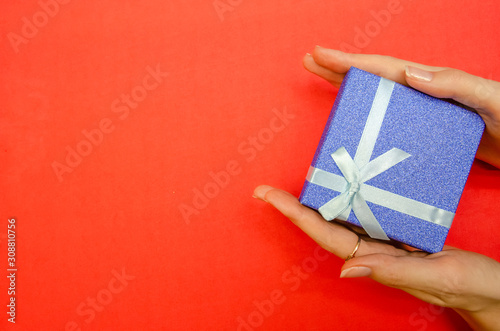Top view of women's hands holding a shiny dark blue gift box with a white bow on a red background, copy space © yaroslav1986