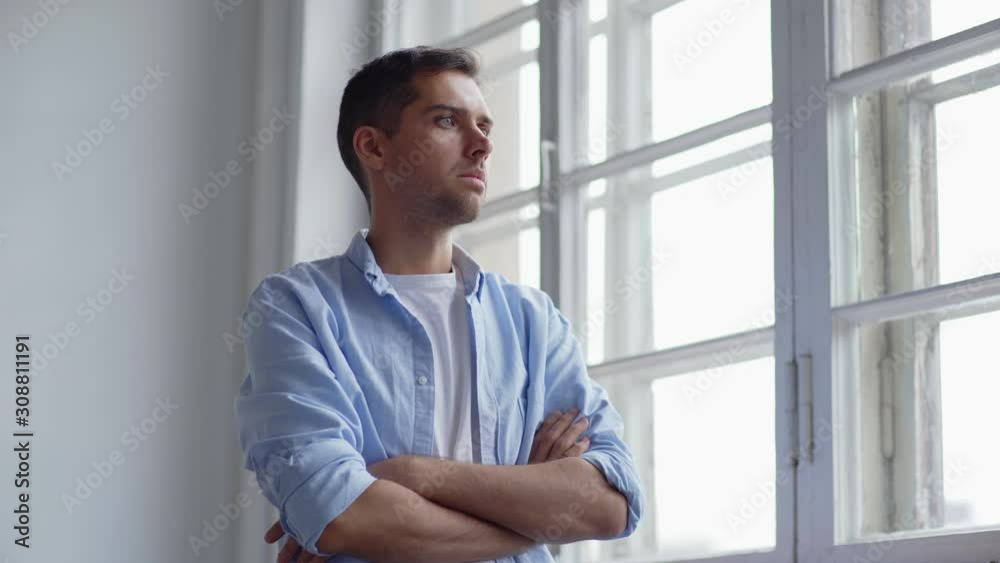 Waist up shot of sad young man standing by window with crossed arms looking through glass and thinking Waist up shot of sad young man standing by window with crossed arms looking through glass and thinking