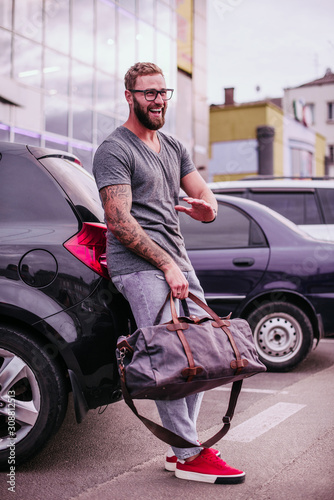 Portrait of young beautiful fashionable man near his car. Hipster style guy with trendy leather bag © VlaDee