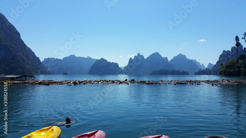 .Beautiful limestone islands and mountain carvings (under a blue sky, with a swimmer and bright colored kayaks) seen from the platform of a remote resort at the Khao Sok National Park, Thailand