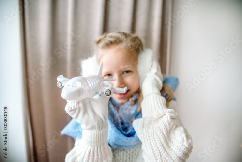 A child plays with an airplane, a cozy winter in a knitted sweater and a white fluffy hat. music in fur headphones. Winter travel concept. Winter holiday. Classic blue scarf