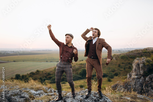Two businessmen farmers celebrating a triumph with arms up. Excited partners jumps up sunset sky, celebrating good teamwork result. Great collaboration. Victory, success and achievement concept.