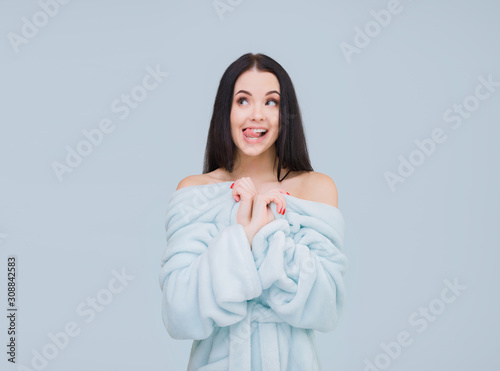 Young sexy girl in bathrobe with bare shoulders posing on gray background. Pretty woman expresses joyful emotions. Free space for text.