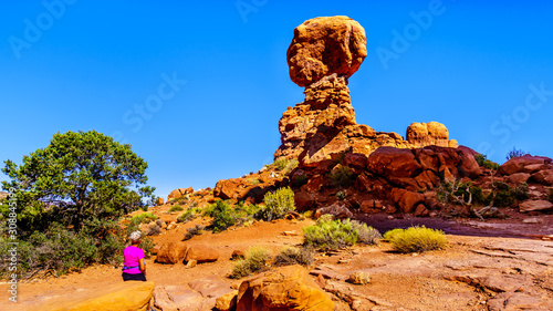 Active Senior Woman on a hike looking at Balanced Rock, a tall and delicate sandstone Rock Formation in the desert landscape of Arches National Park near Moab in Utah, United States photo