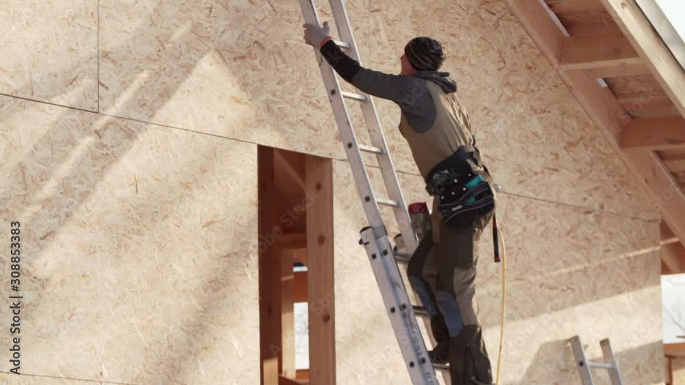 Professional builder is climbing high steel ladder with hydraulic hammer in hand. Frame residential house under construction