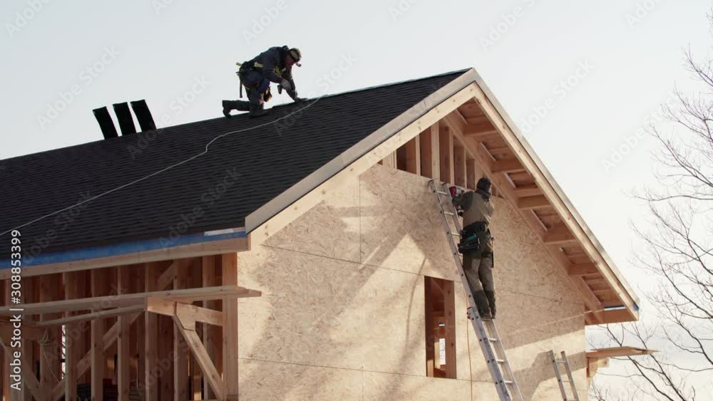 Residential frame house under construction. One worker is covering the wooden frame with plywood using hydraulic hammer, another one is making ridge on the roof in safety harness Residential frame house under construction. One worker is covering the wooden frame with plywood using hydraulic hammer, another one is making ridge on the roof in safety harness