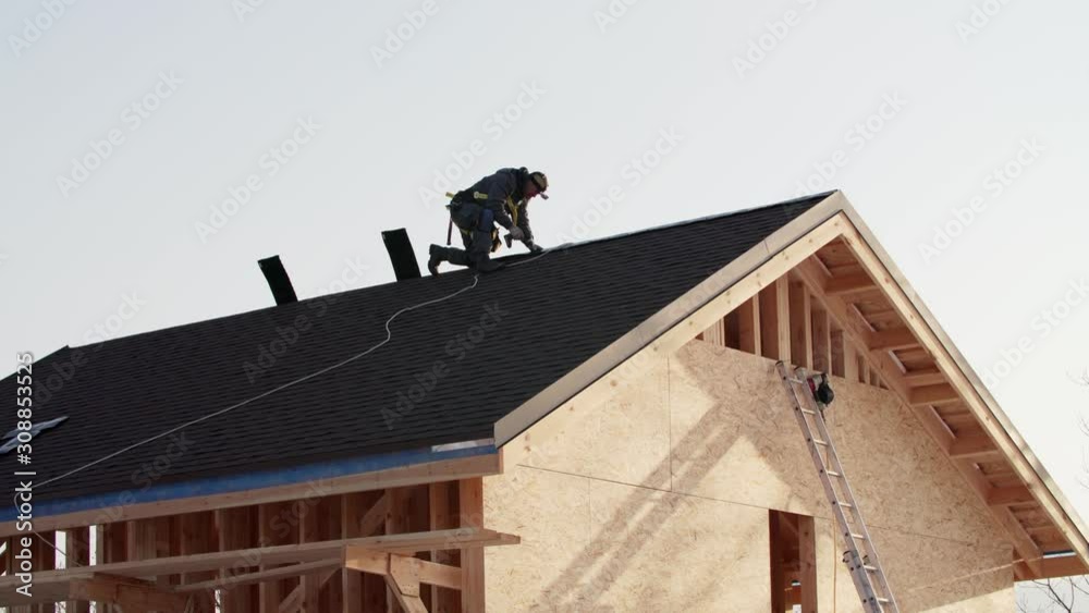 Tiler in safety harness is making roof ridge with hydraulic hammer. Works at height. Wooden frame house under construction Tiler in safety harness is making roof ridge with hydraulic hammer. Works at height. Wooden frame house under construction