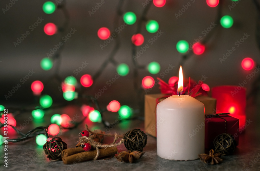 Red and white festive candles burn with a yellow flame on a table among gifts in craft paper, rattan balls, cinnamon sticks and anise stars on the background of a multi-colored Christmas garland